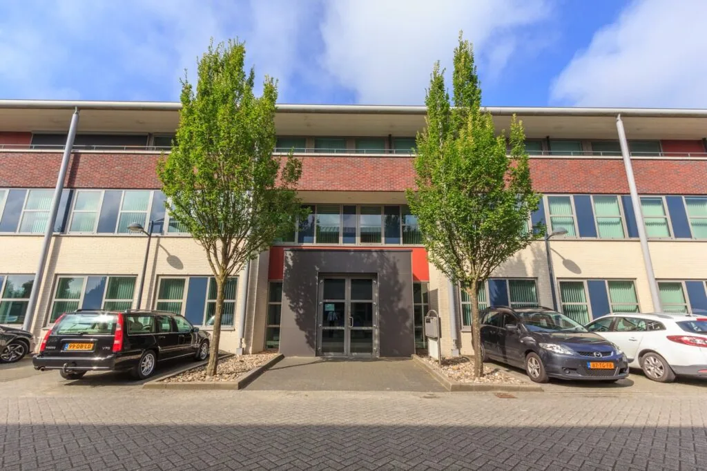 Vooraanzicht van het kantoorgebouw De Wel met bomen en geparkeerde auto's ervoor.